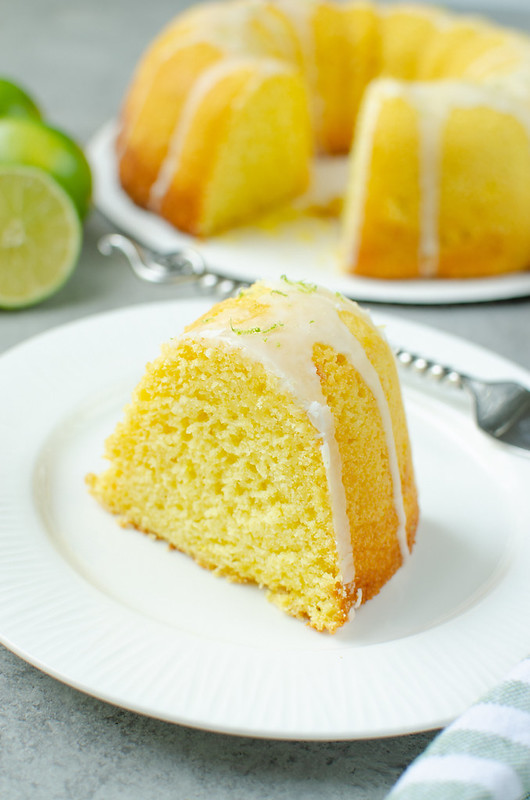 Slice of margarita bundt cake with glaze and lime zest on top; on a white plate with a fork. Limes and the rest of the cake in the background