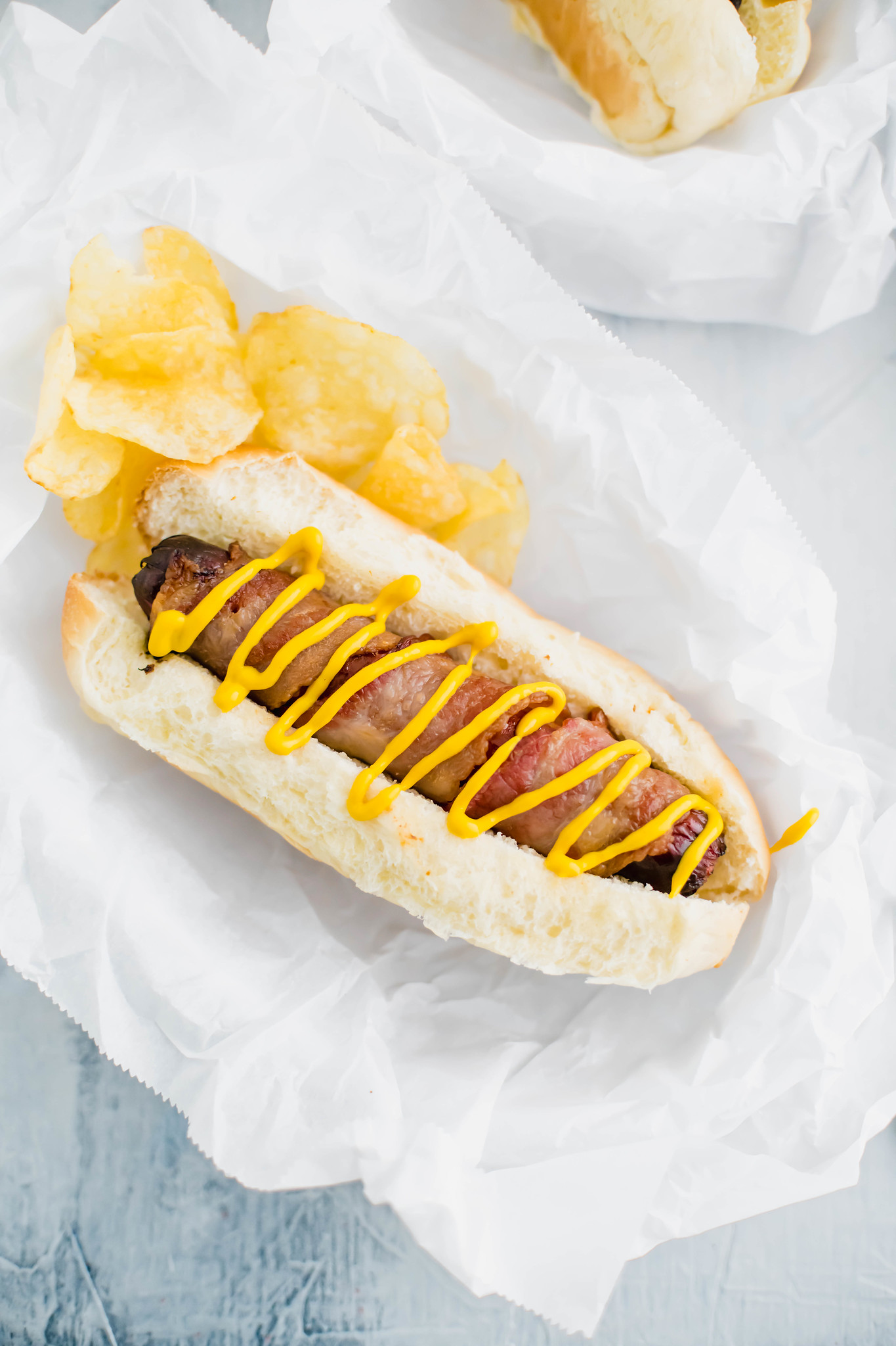 bacon wrapped air fryer hot dog in a bun drizzled with yellow mustard and chips on the side