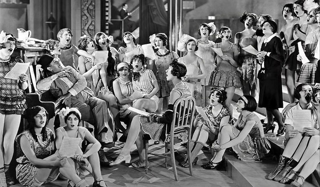 Early 1930s women singing with a hearty gusto, some from memory and others from sheet music. The piano player listens with legs crossed. Oddly enough, most seem to be looking up. What on earth is going on here?