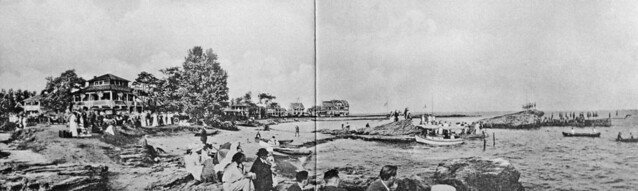 A jolly Fourth of July at the Anchor Beach. Seven flags fly from a house, women wear their finest, men celebrate in suits, people enjoy canoeing, a small covered boat docks and swimmers gather on the Signal Rock and its pier. Milford CT. July 1917