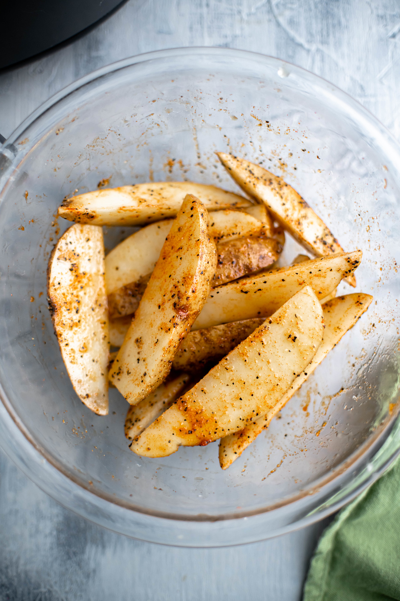 Potato wedges tossed in olive oil and spices in a glass bowl before being cooked.
