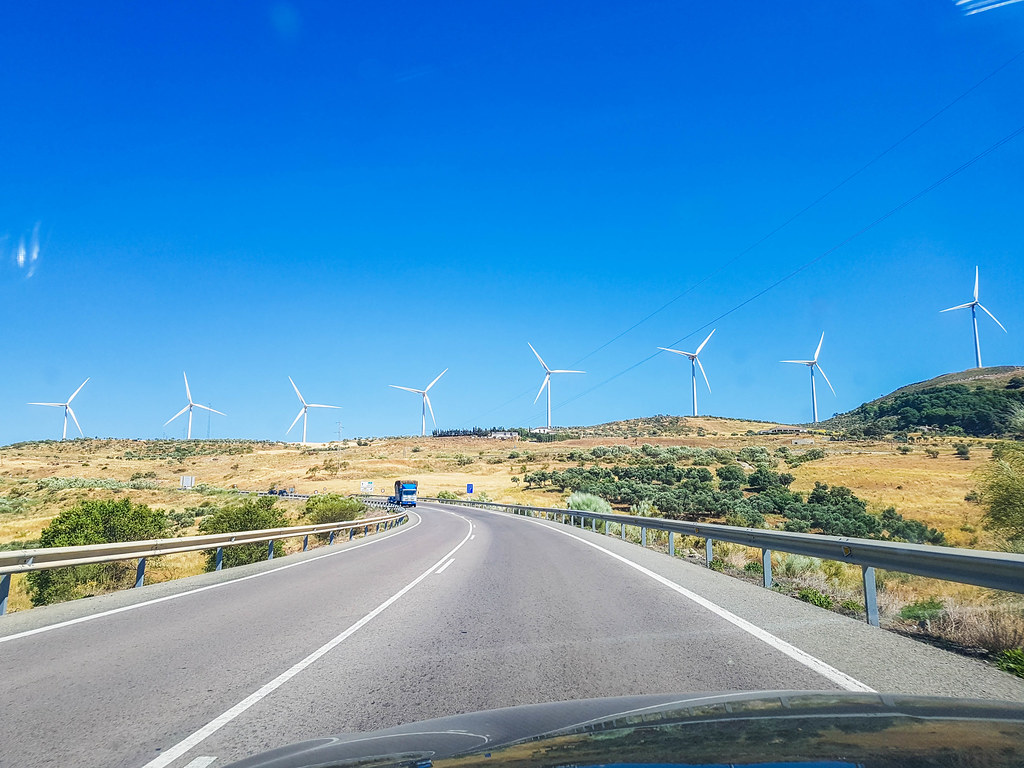 A view of a road, seen through the windscreen on a car. In front there is a hill with white wind turbines on it