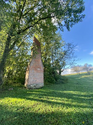 The remains of an old brick chimney stand at the edge of a field with trees growing behind it.