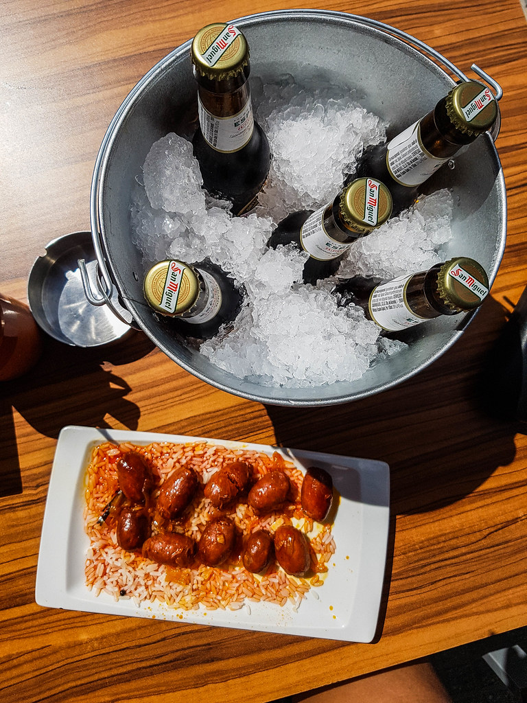 A small bucket with five beers, next to a rectangular plate filled with rice topped with small chorizo sausages