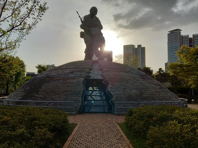 A statue of two soldiers embracing sits atop a small domed structure surrounded by a railing, brick walkway and gardens.