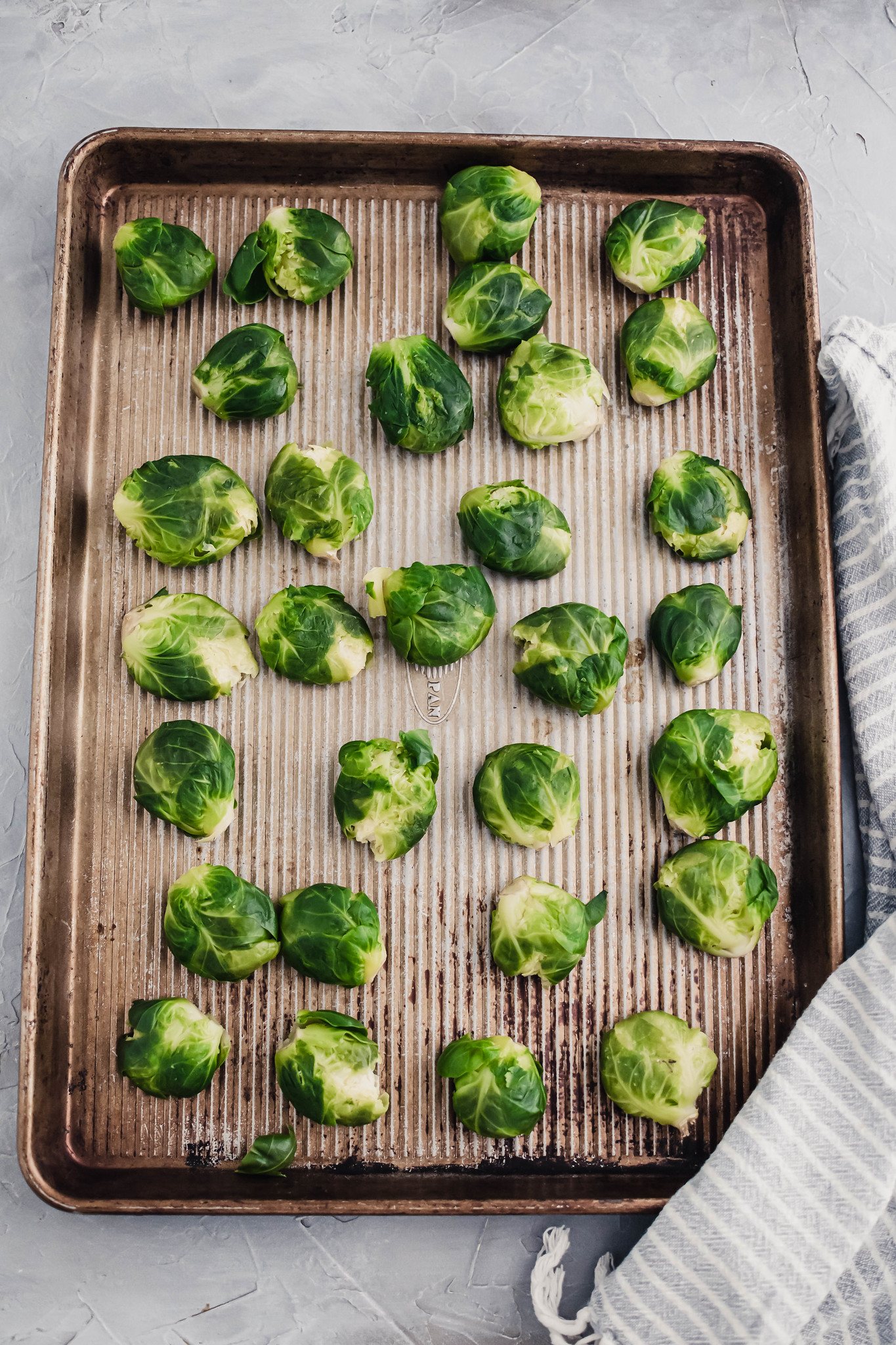 parboiled and smashed brussels sprouts on a baking sheet with a blue and white striped cloth napkin on the right side.