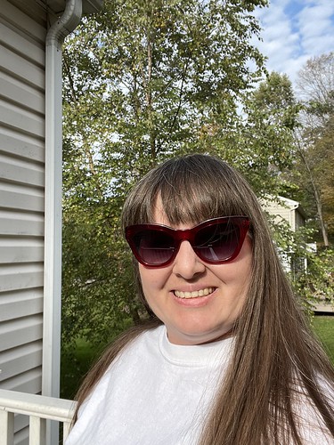 Picture of a female owner of this blog. She is in a white t-shirt, has her hair down, and is wearing red sunglasses. In the background you can see part of a house with tan siding and a white gutter. Behind that is a tall tree in various shades of green. 