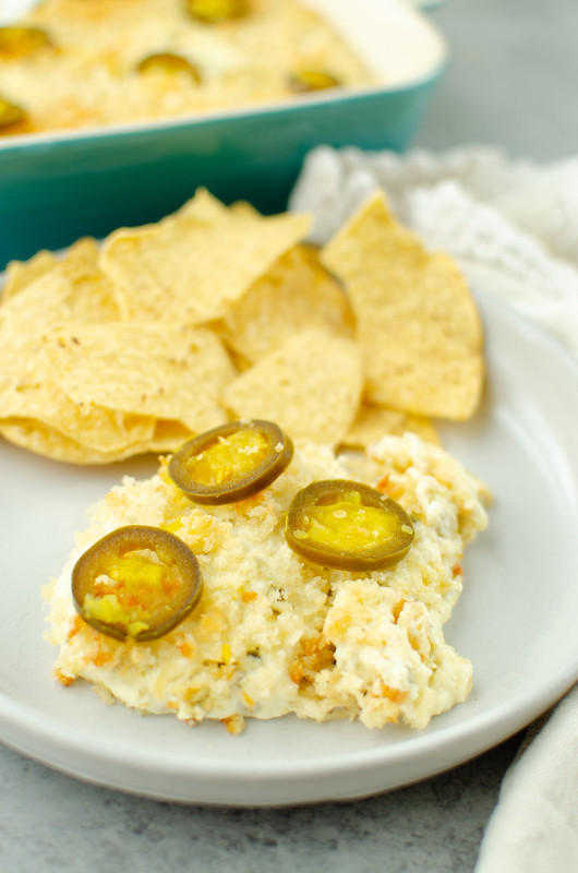 Jalapeno popper dip with jalapenos on top with a white plate and tortilla chips in the background