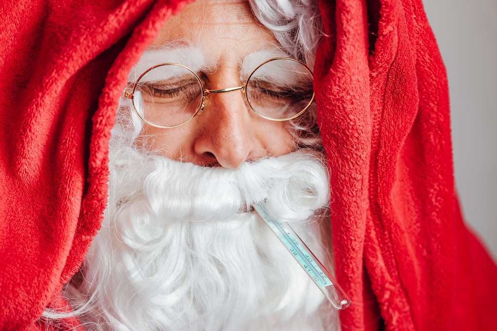 Erkälteter Weihnachtsmann eingewickelt in rote Bettdecke mit Fiebermesser im Mund