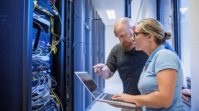 Two people in a server room looking at the configuration and a laptop. 