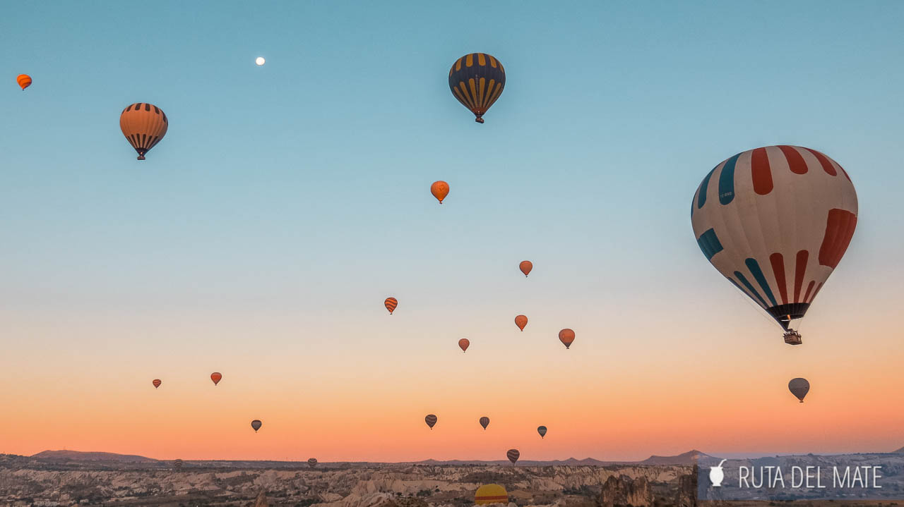 Hot air balloons of different colors floating in the clear sky of Cappadocia. The irregular terrain of Cappadocia is seen, with its sharp karst rocks.