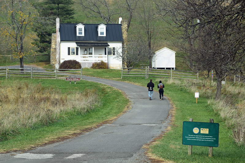 A white house at the end of a paved road, with two people viewed from behind and in the distance walking on the road toward the house.