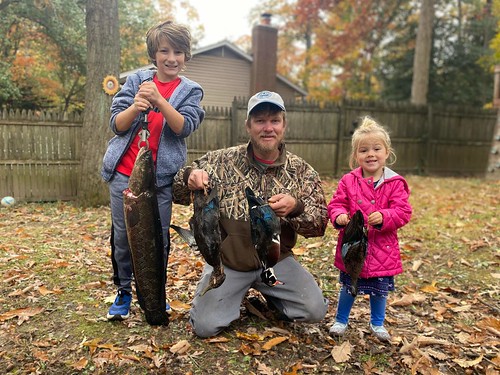 Photo of man with two kids in a yard, holding ducks and a fish that they harvested
