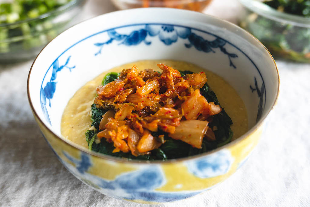 A bowl of yellow hominy topped with sautéed spinach and fried kimchi.