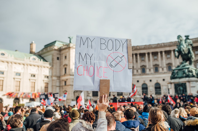 Austrian man protesting vax mandate holding sign with text "My Body My Choice" and an image of a scratched through vaccine