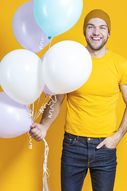 Celebrations Concepts. Funny Smiling Caucasian Young Handsome Man With Colorful Air Balloons Posing in Warm Hat Over Yellow Background.