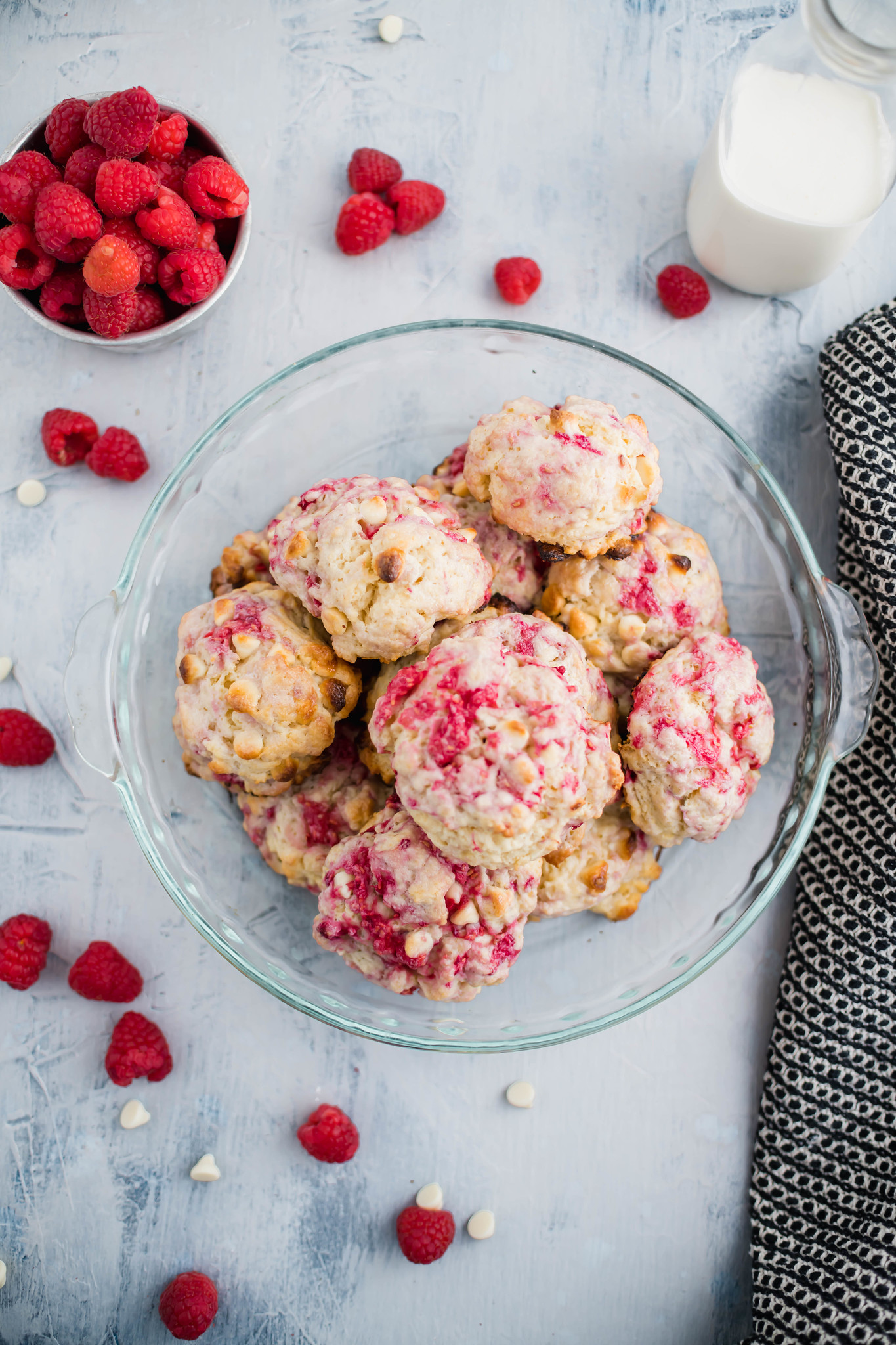 Glass pie plate piled high with raspberry and white chocolate scones. Small bowl of raspberries in upper left corner and heavy cream in a bottle in upper right corner.