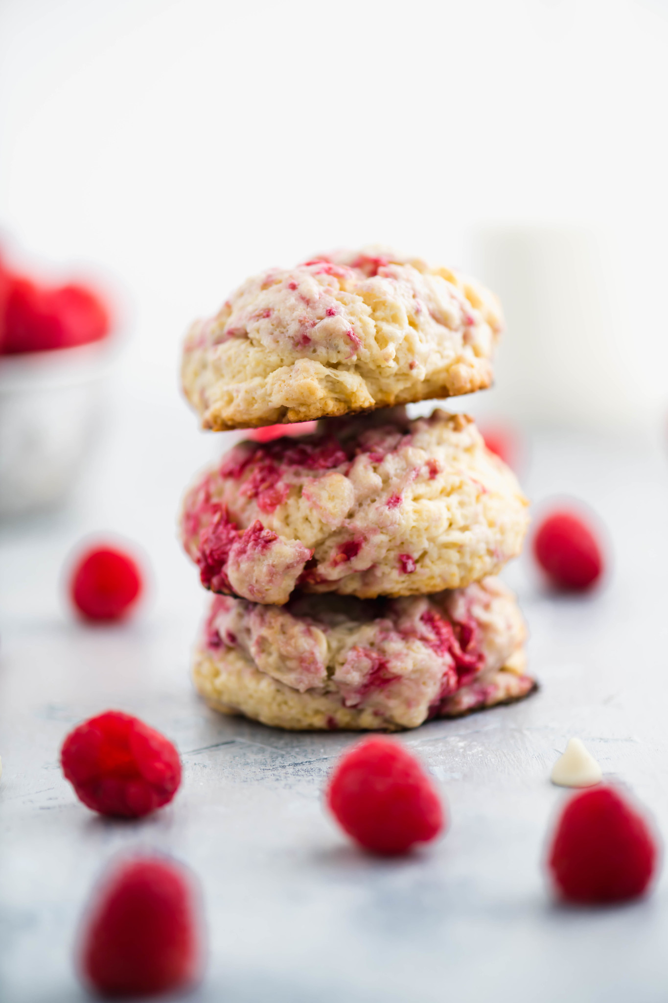 Three raspberry scones stacked on top of each other surrounded by fresh raspberries and white chocolate chips.