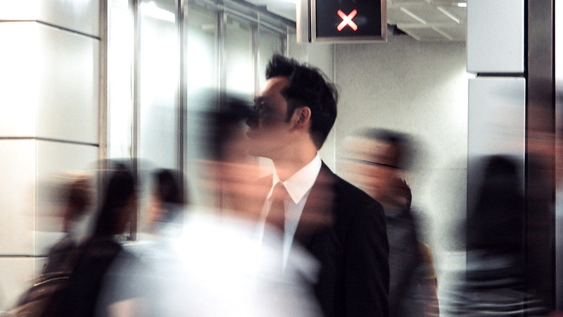 A man wearing a dark suit stood in a busy corridor with other people moving around him.