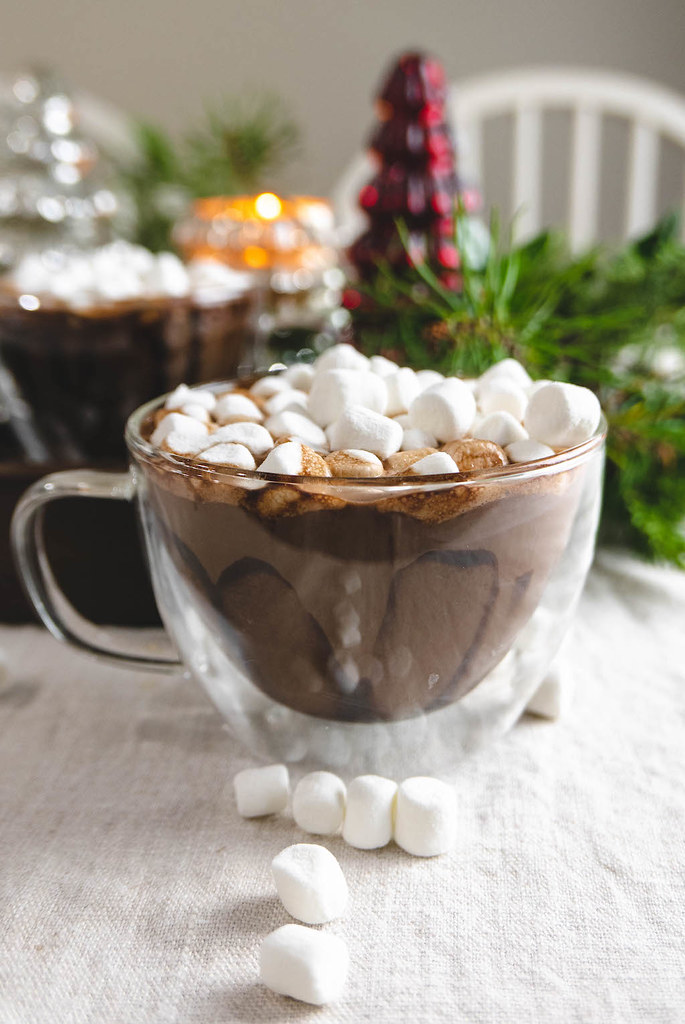 A glass mug of hot chocolate filled with marshmallows. Greenery sits in the back.