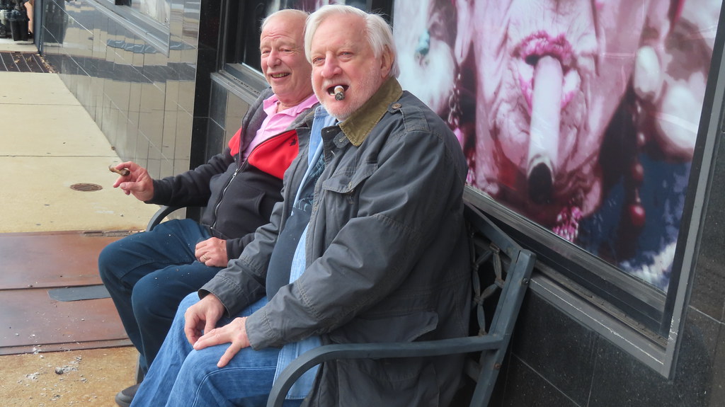 Two elderly gents enjoy cigars outdoors. Perhaps, their wives won't let them smoke at home.