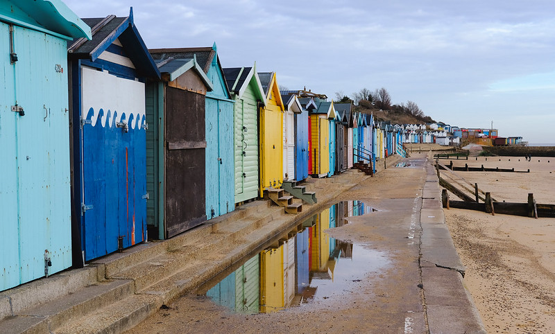 A photo of a row of colourful beach huts receding into the distance, a few of them reflected in a puddle on the concrete promenade.