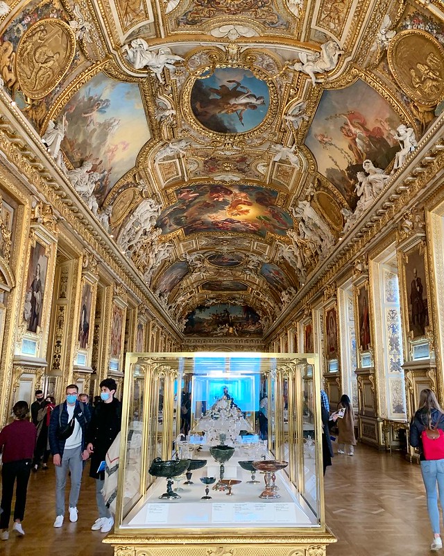 Intricate gold framing lines the ceiling surrounding various works of art. The long hall decorated with paintings also presents art pieces in a glass case.