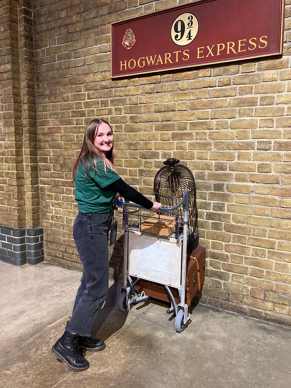 Macy pushing a cart in to the brick wall with the sign for platform 9 and 3/4 for the Hogwarts Express.