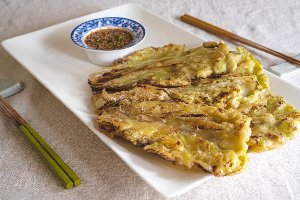 A side shot of Korean cabbage pancakes on a white platter with a dipping sauce in a blue and white bowl. Chopsticks rest next to the tray