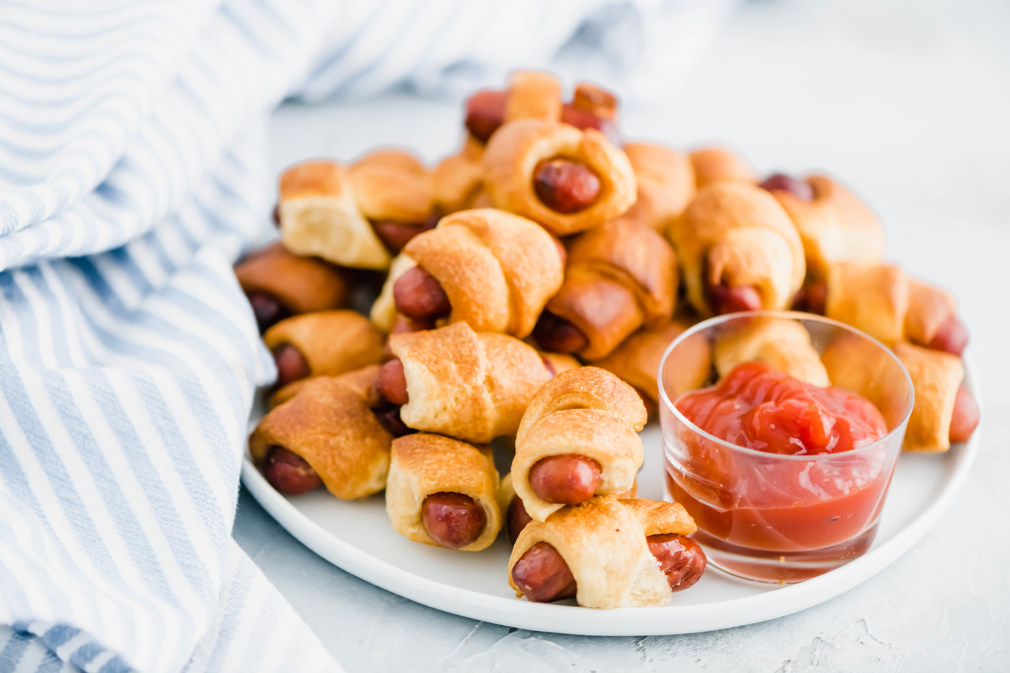 Pile of pigs in a blanket on a white round plate with a glass bowl of ketchup.