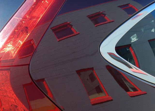 red windows in a blue building reflected in a shiny black car with red taillights