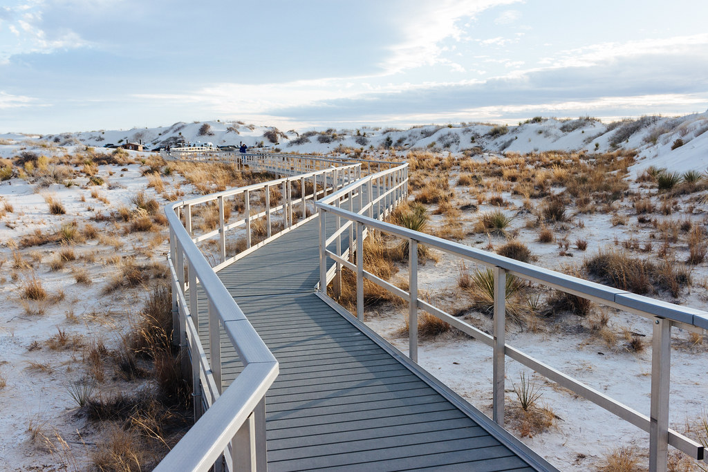 A boardwalk with white railings zig zags through a valley of grass and yucca between two white sand dunes on a late afternoon