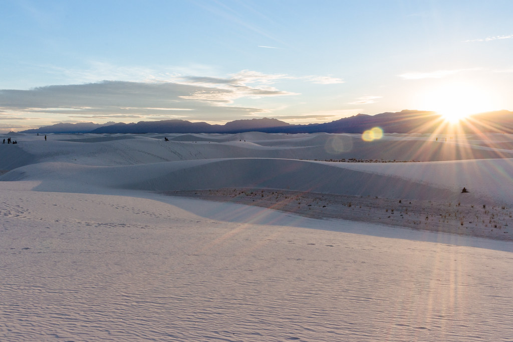 The sun is about to pass beneath purple mountains at sunset over shadowy dunes of white sands
