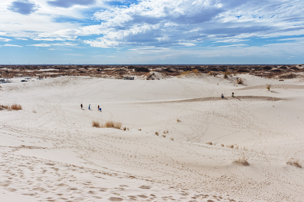 Five people stroll among sand dunes dotted with clumps of grass on a partly cloudy day