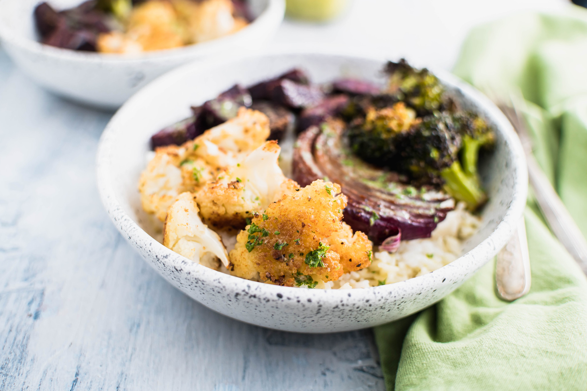 White bowl speckled with black paint. White rice in the base of the bowl topped with roasted cauliflower, roasted broccoli, purple sweet potatoes and roasted onion.