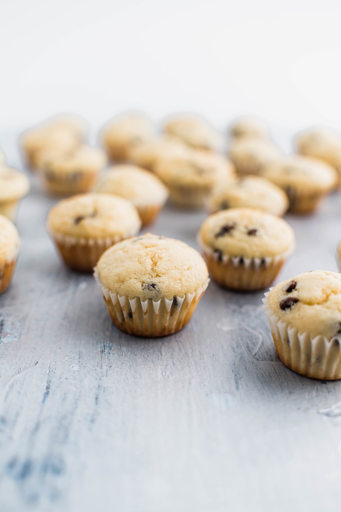 Mini chocolate chip muffins scattered along a gray blue surface.
