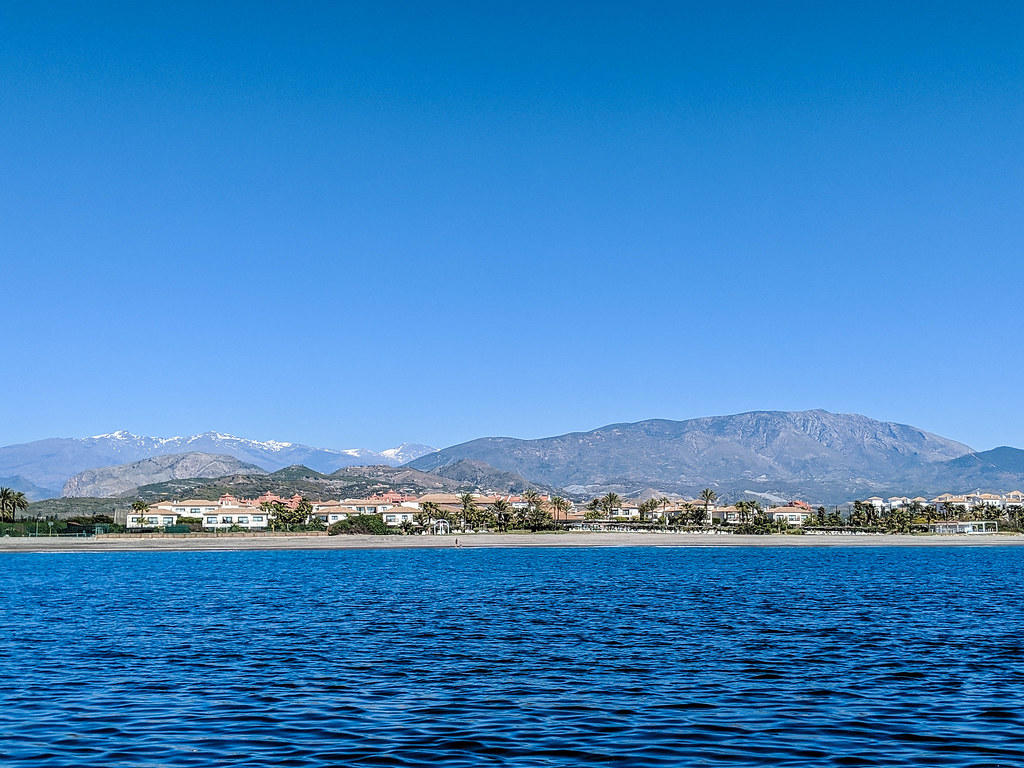 A photo of the beach from afar, taken from a boat. Behind the beach you can see the snowy peaks of the Sierra Nevada mountains.