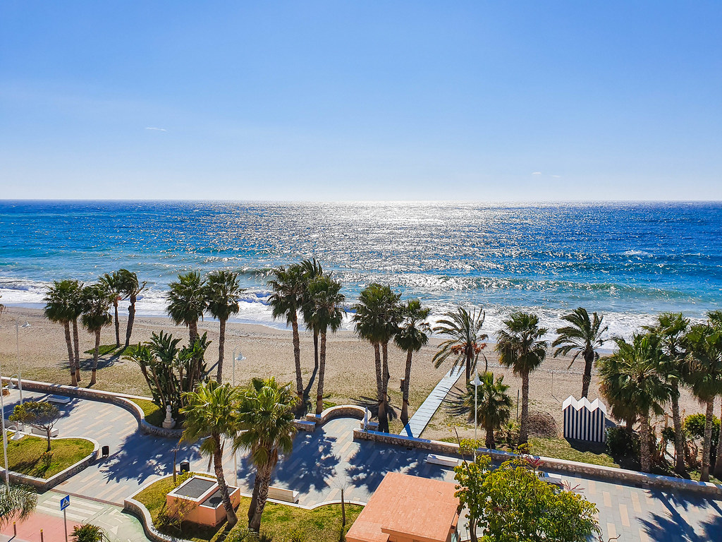 This photo has been taken from the hotel in front. The sun in shining into the sea, making it sparkly. The beach is flanked by tall palm trees, behind which there is a wide paved promenade.