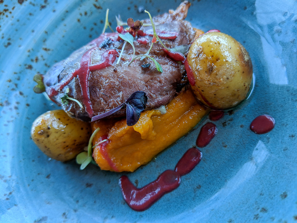 An artistic plating of a meat dish. The plate is blue. The meat is sat on an orange pumpkin puree, with two roast potatoes on each side. the dish is decorated with micro greens and sploshes of red sauce