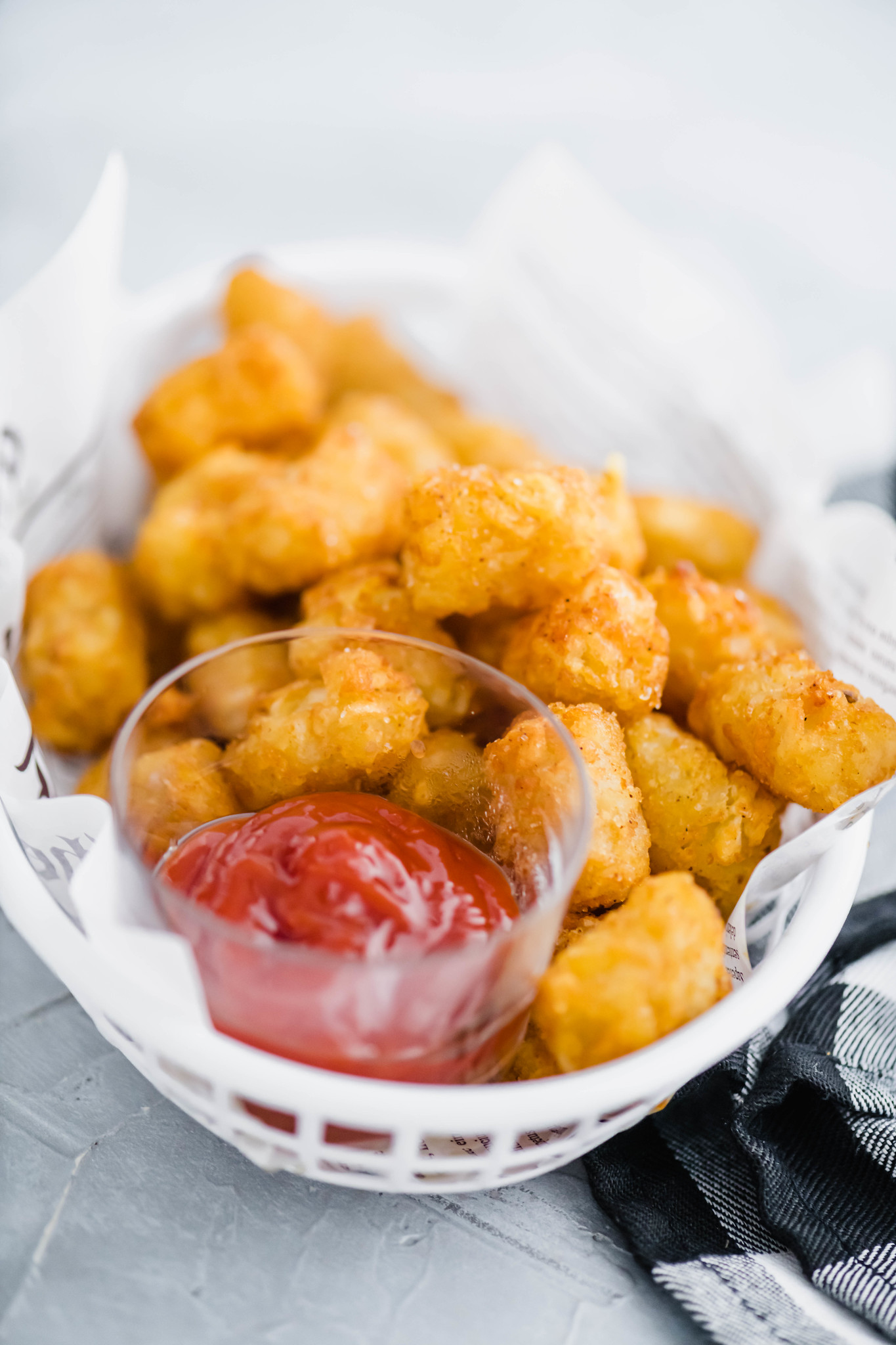 Tater tots in a white plastic fast food basket along with a small glass bowl of ketchup.