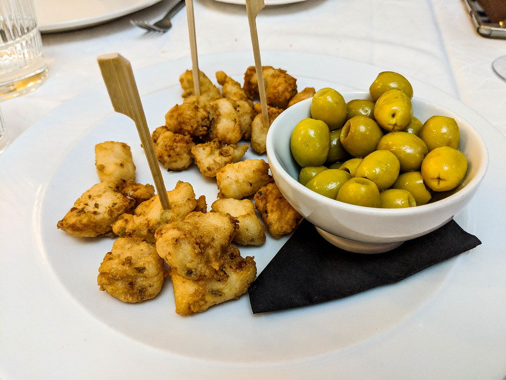 A round plate with small pieces of friend fish, some of which have small wooden skewers in them. Next to the fish, on the same plate, there is a white bowl filled with green olives. Under the bowl there is a black napkin
