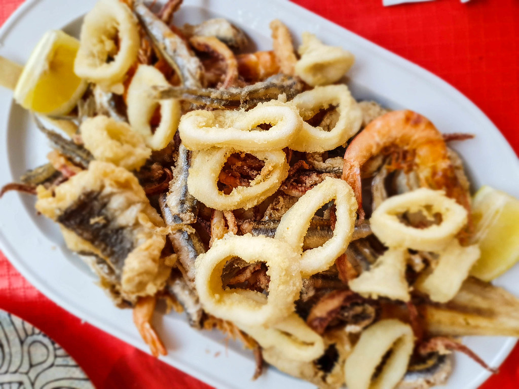 A white round platter filled with different friend fish: small anchovies, squid rings, prawns with the shell on. On each side of the platter there is a wedge of lemon.