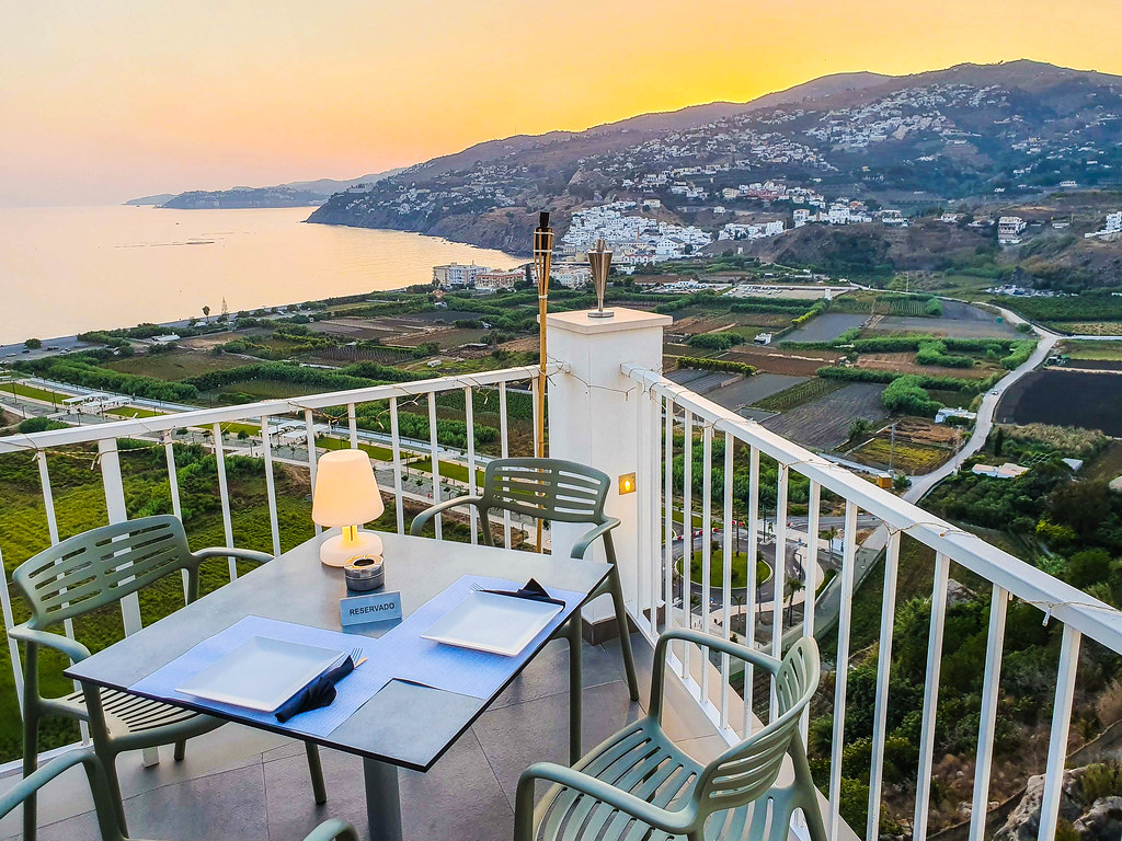 A table at the edge of the terrace, overlooking the valley below. The sea can be seen on the left hand side of the photo. The sky is orange as it is sunset time.