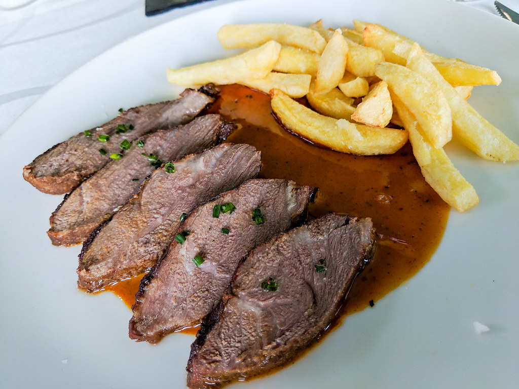 Five slices of meat plated next to each other on a white plate, on a brown sauce. Near the slices of meat there are a few thick cut French fries. 