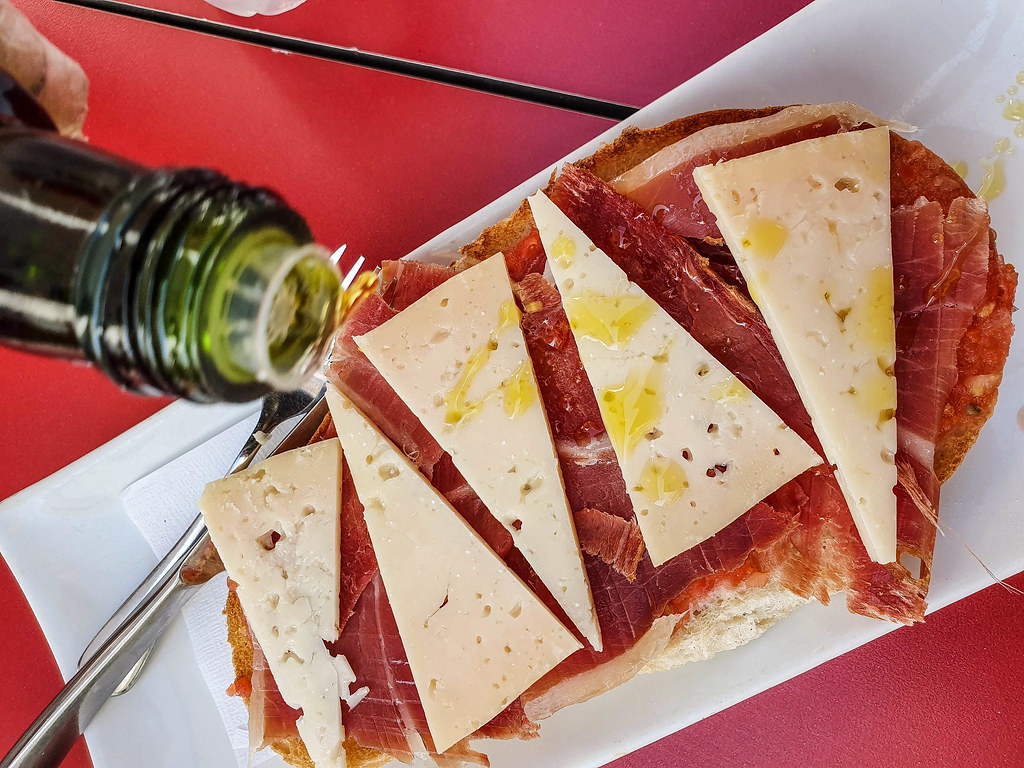 A slice of bread covered with red jamon and sliced of yellow hard cheese. The cheese has olive oil sprinkled over. There is a hand holding a bottle of olive oil above the slice of bread. The bread is on a white, rectangular plate.