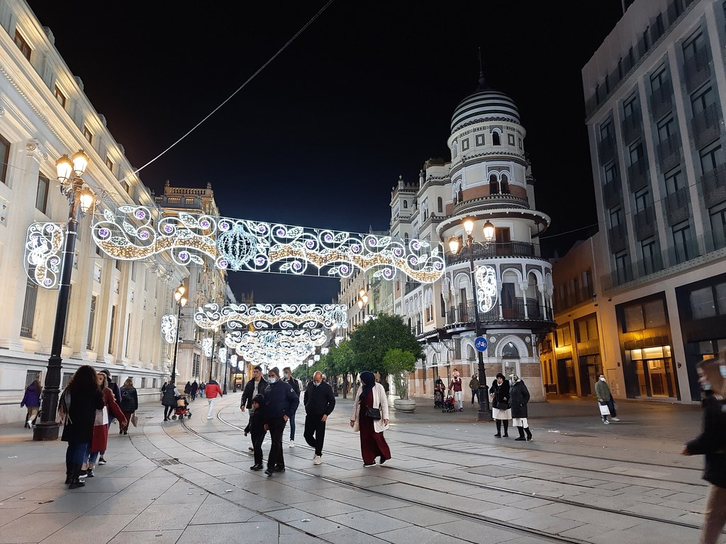 A main pedestrian boulevard in Seville, at night, lit up with white lights. There are people walking around, in all directions. Above the street, there are sets of lights depicting Christmas decorations such as bells and leaves