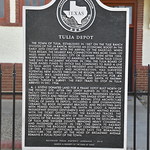 Old Santa Fe Passenger Depot (Tulia, Texas) Historical marker for the Old Santa Fe Passenger Station in Tulia, Texas.  The marker reads:

“The town of Tulia, established in 1887 on the Tule Ranch division of the Ja ranch, received an economic boost in the early 20th century with the arrival of the railroad. When Tulia began, the nearest rail connection was more than 100 miles away in Colorado City or Quanah. Even after 1888, when rail was extended to Amarillo, a trip from Tulia could take days in inclement weather. In 1906, the Tulia Board of Trade raised funds to entice rail companies to build a line to Tulia. Avery Turner, vice-president and general manager of the Pecos Valley and Northeastern, had surveyed potential new routes through the region, and in Jan. 1906 grading was underway south from Canyon for an extension of the Atchison, Topeka and Santa Fe Railway along Turner’s survey. The first train to stop in Tulia arrived in Dec. 1906. A. J. Bivens donated land for a frame depot built north of the present site. After this depot burned in 1915, a new facility was built in 1916-17, combining a passenger station, express office and freight house. This single-story brick and stucco building exhibits mission revival style architecture typical of Santa Fe depots, including a deep overhanging ceramic tile roof, prominent brackets, and projecting bays with peaked parapets and the Santa Fe logo. A narrow passageway divided gentlemen’s and ladies’ waiting rooms, with the ticket office facing the railroad tracks. The baggage room was north of the passenger section. The coming of the railroad was a milestone in the development of Tulia and Swisher County, bringing heavy commercial and passenger activity. In 1987, the Santa Fe railway demolished the baggage area, but concerned citizens and Swisher County officials helped save the remaining structure. The depot at the head of Broadway Avenue remains a focal point of the town.”