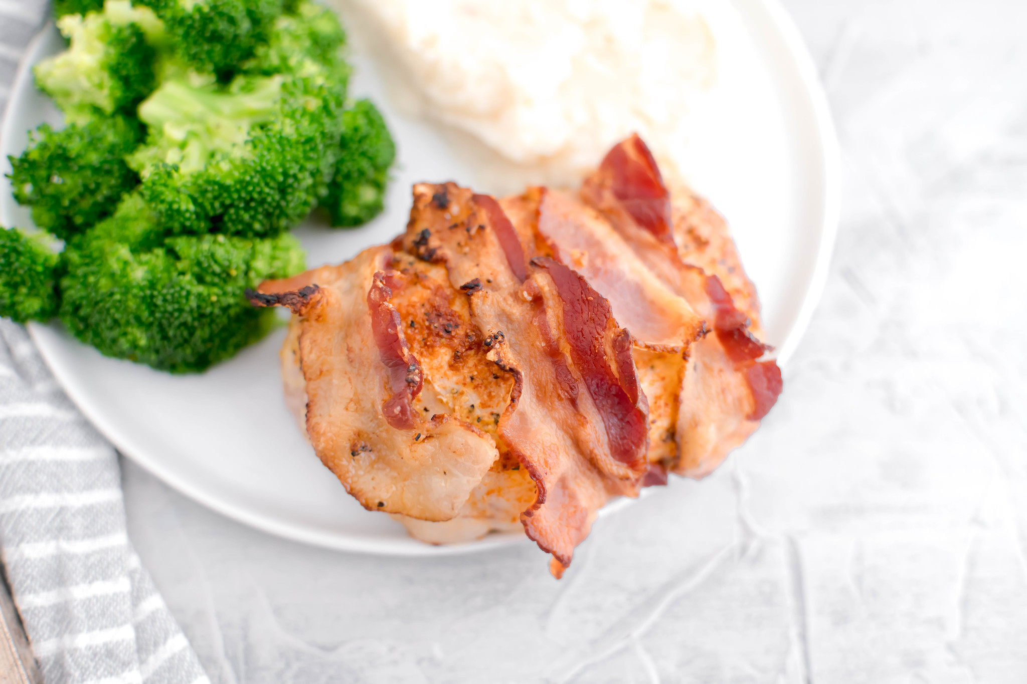 Close up of bacon wrapped pork chop on a round white plate with broccoli and mashed potatoes.