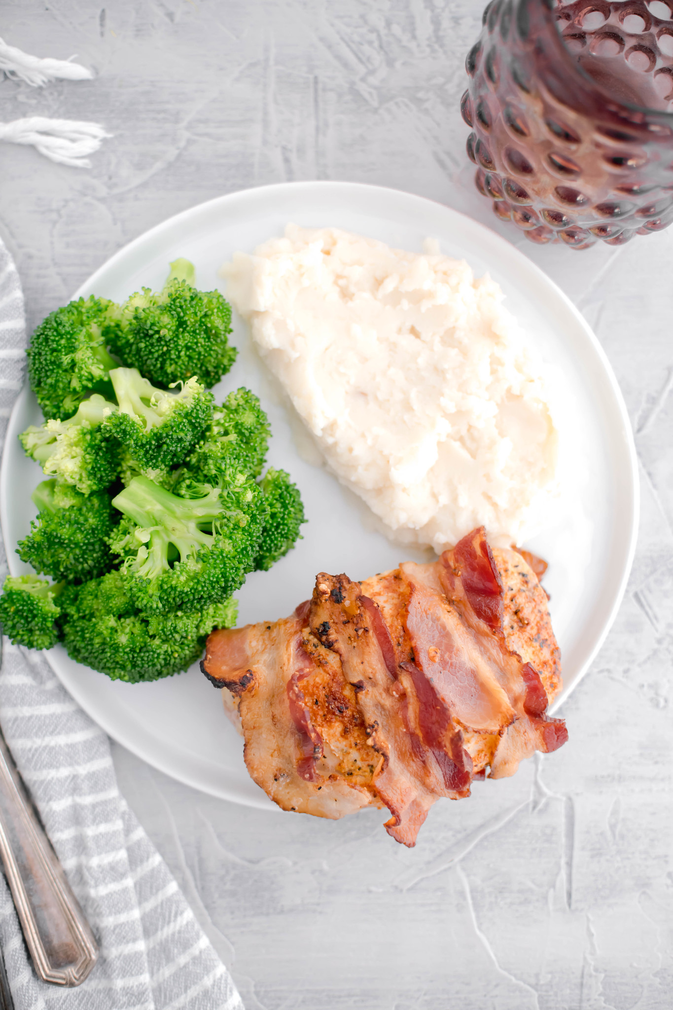Round white plate filled with bacon wrapped pork chop, steamed broccoli and mashed potatoes.
