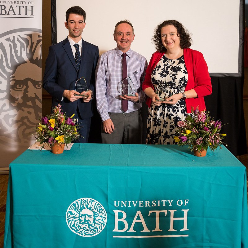 Three teachers stood up behind a table with a teal blue University of Bath logo table cloth, receiving their awards
